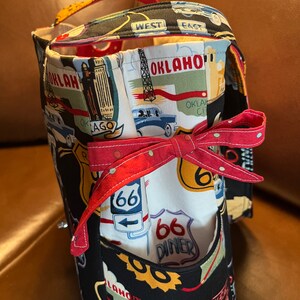 May include: Black fabric tote bag with a Route 66 theme. The bag features a red, white, and blue design with images of cars, diners, and signs. The bag has a red ribbon tied around it.