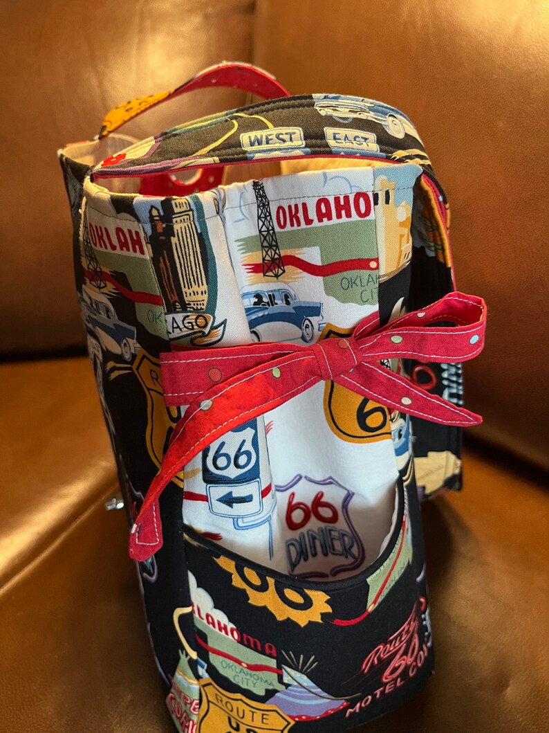 May include: Black fabric tote bag with a Route 66 theme. The bag features a red, white, and blue design with images of cars, diners, and signs. The bag has a red ribbon tied around it.