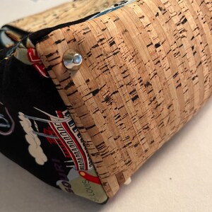 May include: A close-up of a cork fabric bag with a metal rivet. The bag is made of a black fabric with a colorful pattern of a city skyline and the words "St. Louis" and "Missouri".
