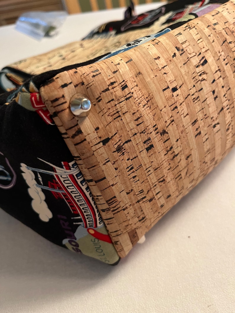 May include: A close-up of a cork fabric bag with a metal rivet. The bag is made of a black fabric with a colorful pattern of a city skyline and the words "St. Louis" and "Missouri".