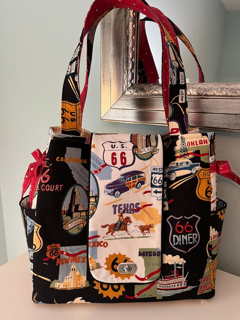 May include: A black tote bag with a Route 66 theme. The bag features a variety of images and text related to the iconic highway, including a vintage car, a diner sign, and a cowboy riding a horse. The bag has red and white polka dot straps.