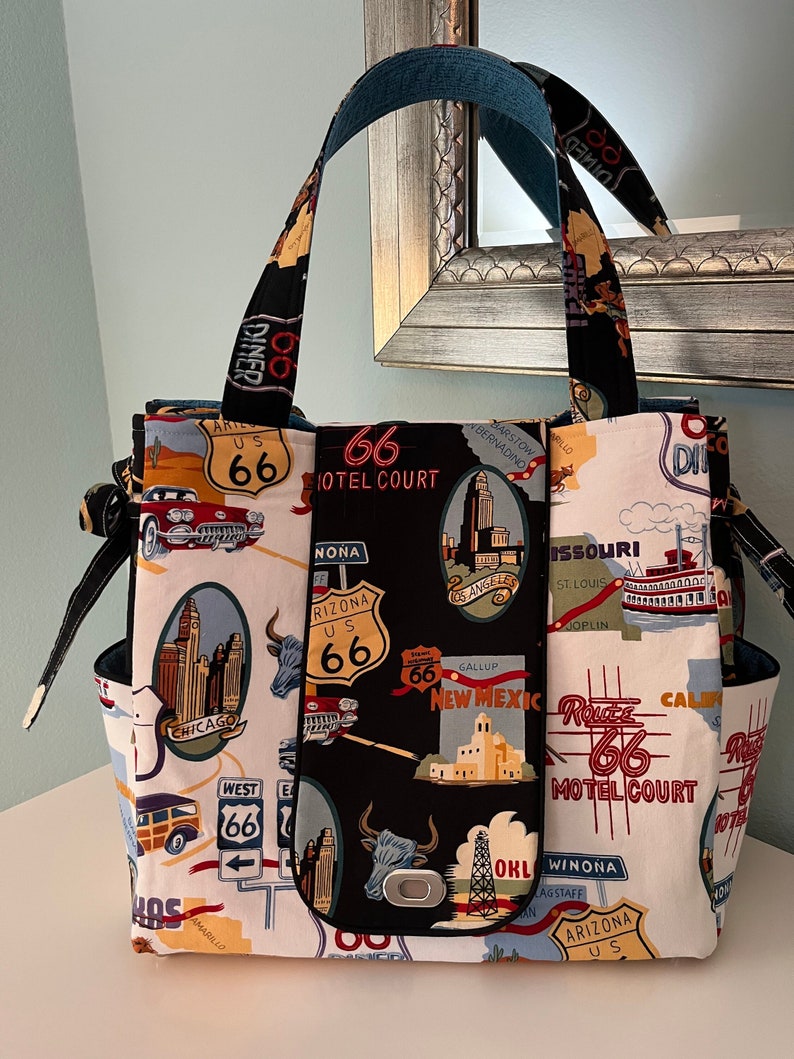 May include: A black and white tote bag with a Route 66 theme. The bag features a variety of images and text related to Route 66, including a motel sign, a car, and a map. The bag has a black handle and a black zipper.
