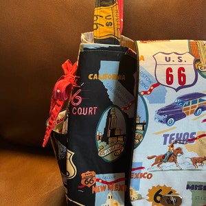 May include: A black and white fabric tote bag with a Route 66 theme. The bag features images of iconic landmarks, including the Los Angeles skyline, a cowboy riding a horse, and a vintage car. The bag has a red strap and a silver clasp.