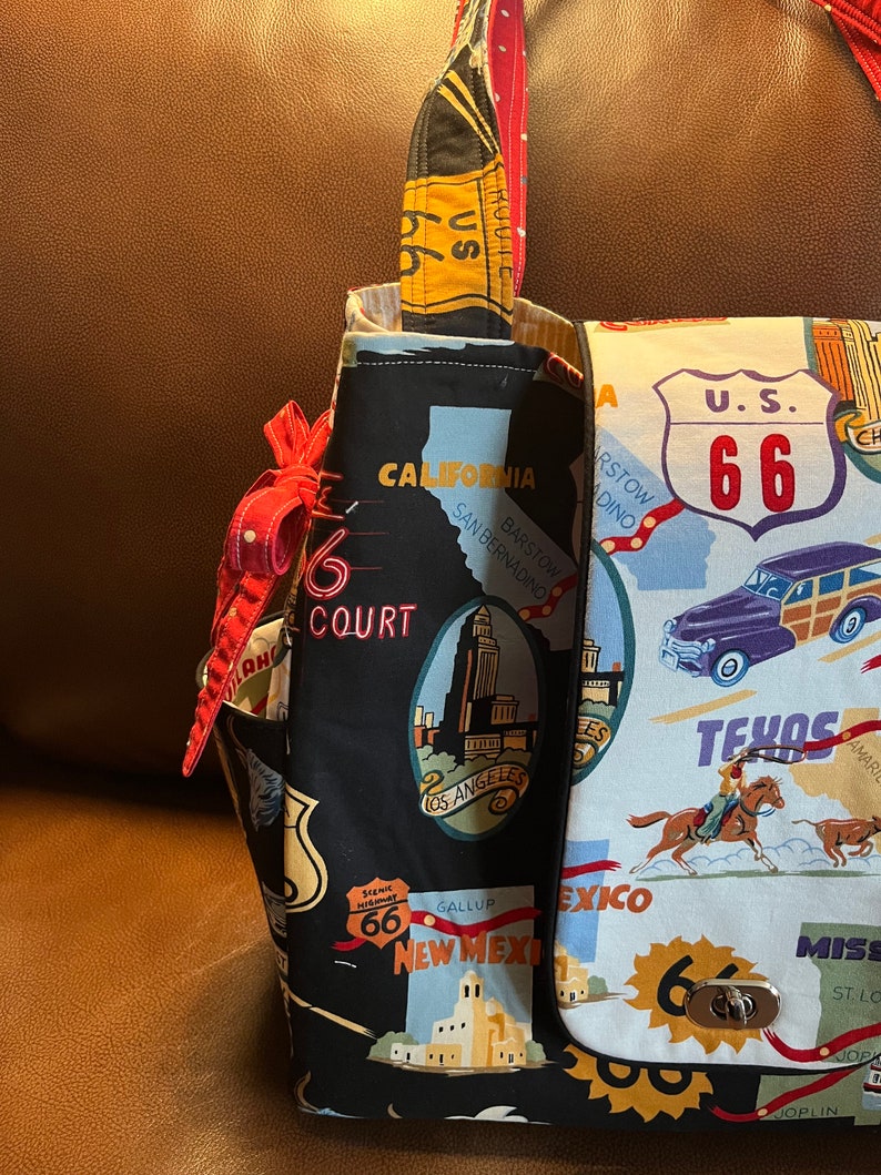 May include: A black and white fabric tote bag with a Route 66 theme. The bag features images of iconic landmarks, including the Los Angeles skyline, a cowboy riding a horse, and a vintage car. The bag has a red strap and a silver clasp.
