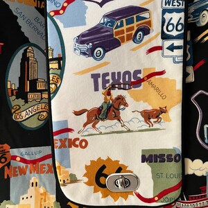 May include: A black fabric bag with a white fabric pocket featuring a Route 66 theme. The pocket has a metal clasp and is decorated with images of a car, a cowboy riding a horse, and a cow. The text "Texas", "Amarillo", "Mexico", "Gallup", "New Mex", "Missou", "St. Louis", and "Joplin" is printed on the pocket.