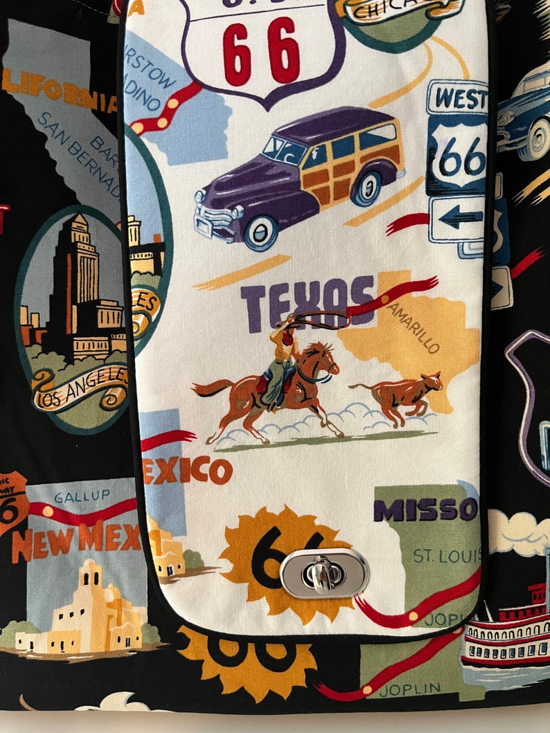 May include: A black fabric bag with a white fabric pocket featuring a Route 66 theme. The pocket has a metal clasp and is decorated with images of a car, a cowboy riding a horse, and a cow. The text "Texas", "Amarillo", "Mexico", "Gallup", "New Mex", "Missou", "St. Louis", and "Joplin" is printed on the pocket.