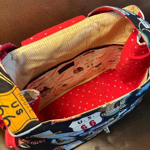 May include: A black fabric tote bag with a red and white polka dot lining. The exterior of the bag features a vintage travel theme with images of road signs, cars, and state names. The bag has a zipper closure and two red straps.
