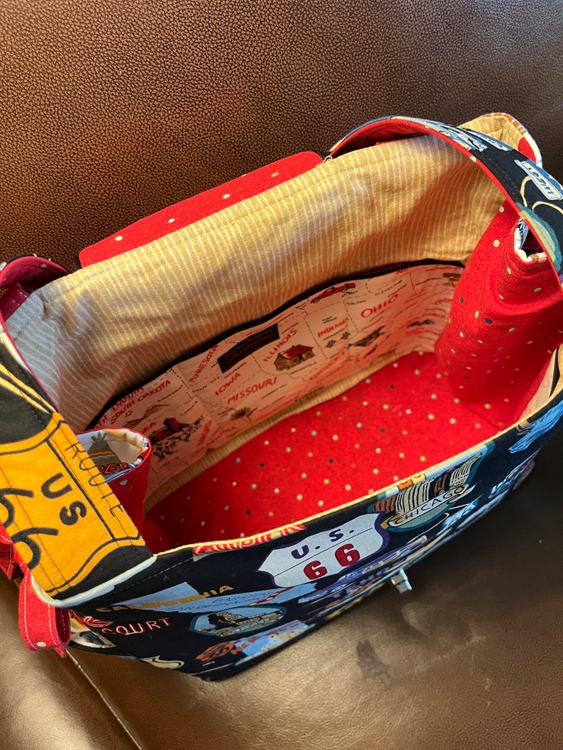 May include: A black fabric tote bag with a red and white polka dot lining. The exterior of the bag features a vintage travel theme with images of road signs, cars, and state names. The bag has a zipper closure and two red straps.