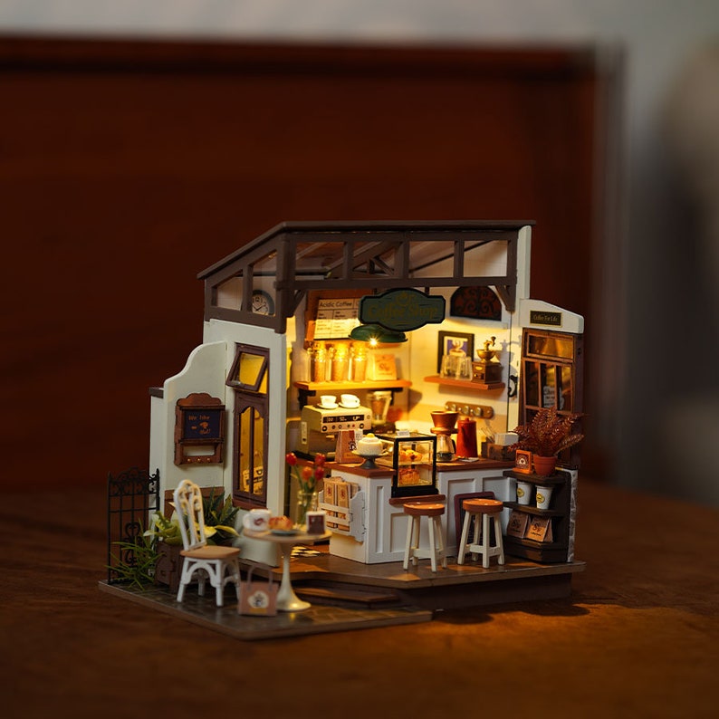 May include: Miniature coffee shop model with a detailed interior. The model features a counter with stools, a coffee machine, and shelves with coffee-related items. The exterior has a small table and chairs, and a sign that reads "Acide Coffee".