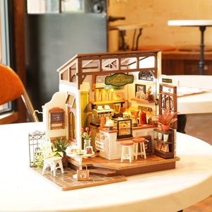 May include: Miniature coffee shop model with a detailed interior and exterior. The model features a sign that reads "Coffee Shop" and includes miniature furniture, decor, and a small table with a chair. The model is made of wood and has a warm, inviting aesthetic.