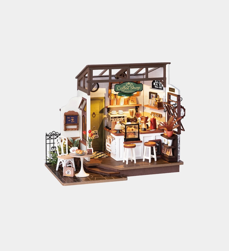 May include: Miniature coffee shop model with a green and white sign that reads "Coffee Shop". The model includes a small table and chairs, a counter with stools, and various coffee-making equipment. The shop has a rustic, inviting aesthetic.
