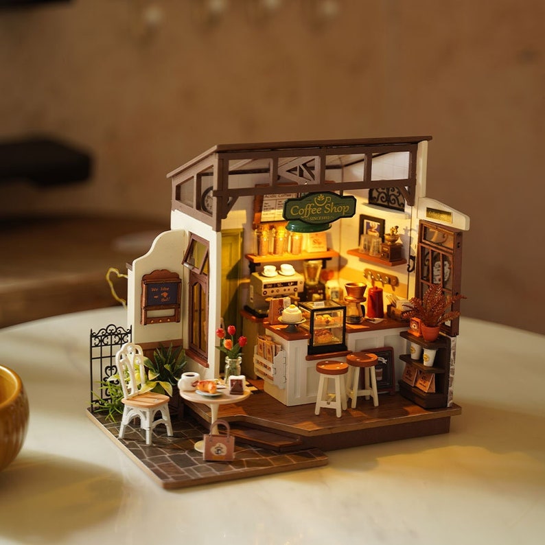 May include: Miniature coffee shop model with a detailed interior. Features a counter with stools, a coffee machine, and shelves with coffee-related items. Includes a small table with a chair and a sign that reads "Coffee Shop".