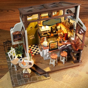 May include: Miniature coffee shop model with a detailed interior. Features a counter with stools, a coffee machine, and a display case with pastries. Includes a small table with a chair, coffee, and a pastry. The sign reads "Coffee Shop".
