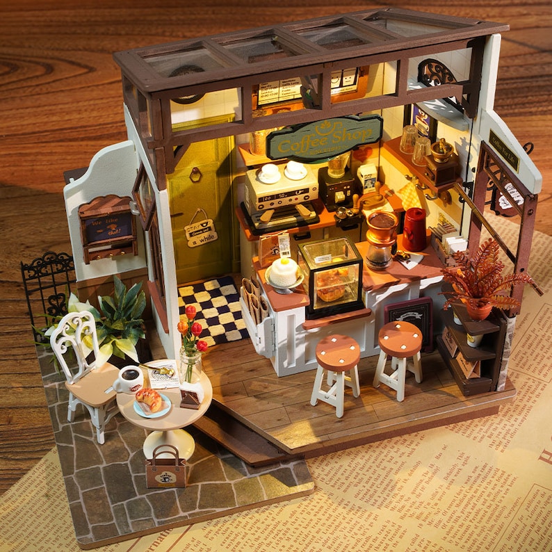 May include: Miniature coffee shop model with a detailed interior. Features a counter with stools, a coffee machine, and a display case with pastries. Includes a small table with a chair, coffee, and a pastry. The sign reads "Coffee Shop".