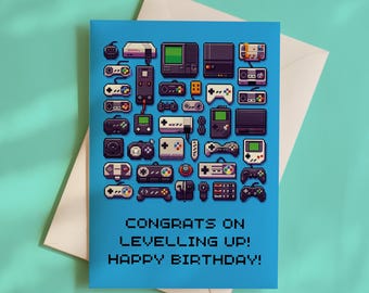 May include: A blue greeting card featuring a pixel art collage of various vintage video game consoles and controllers. The text at the bottom reads: CONGRATS ON LEVELLING UP! HAPPY BIRTHDAY! The card is placed on top of a white envelope.