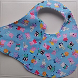 May include: A blue baby bib with a white trim and a snap closure. The bib features a repeating pattern of cartoon pigs in various outfits on a light blue background.