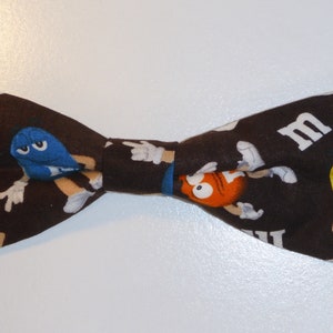 May include: A brown bow tie featuring a pattern of colourful M&M's characters. The bow tie has a white "M" on one side.