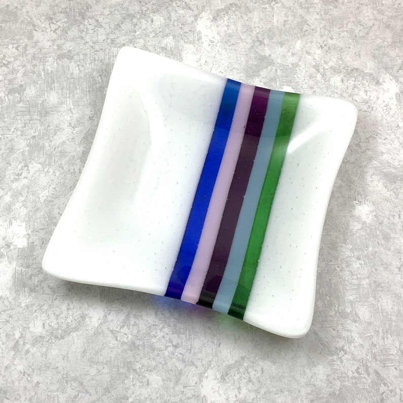 May include: A white square dish with a decorative stripe of blue, green, purple, and pink glass.
