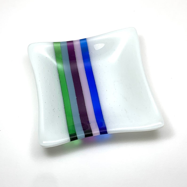 May include: A small, white square glass dish with a single vertical stripe of green, blue, purple, and pink glass.