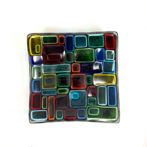 Puede incluir: Un plato cuadrado de vidrio con un dise&ntilde;o de mosaico. El plato est&aacute; hecho de peque&ntilde;os cuadrados de vidrio en varios colores, incluyendo rojo, verde, azul, amarillo y negro. El plato tiene un borde negro.