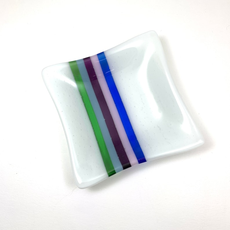 May include: A small, white square dish with a decorative stripe of blue, green, purple, and pink glass.