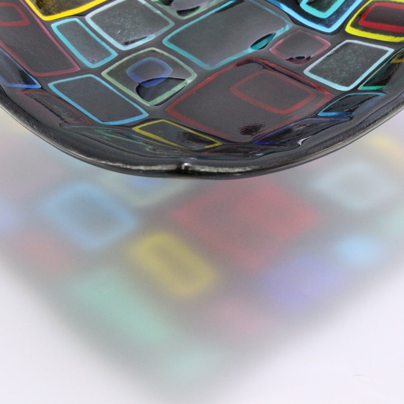 Puede incluir: Un cuenco de vidrio negro con un dise&ntilde;o de mosaico de cuadrados coloridos. El cuenco es ligeramente curvo y tiene una superficie reflectante.