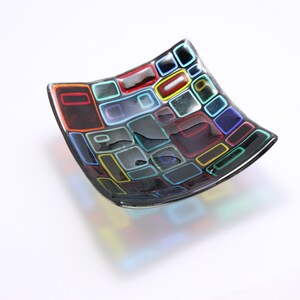 Puede incluir: Un plato cuadrado de vidrio con fondo negro y dise&ntilde;os geom&eacute;tricos coloridos. El plato est&aacute; hecho de vidrio y tiene un acabado brillante.