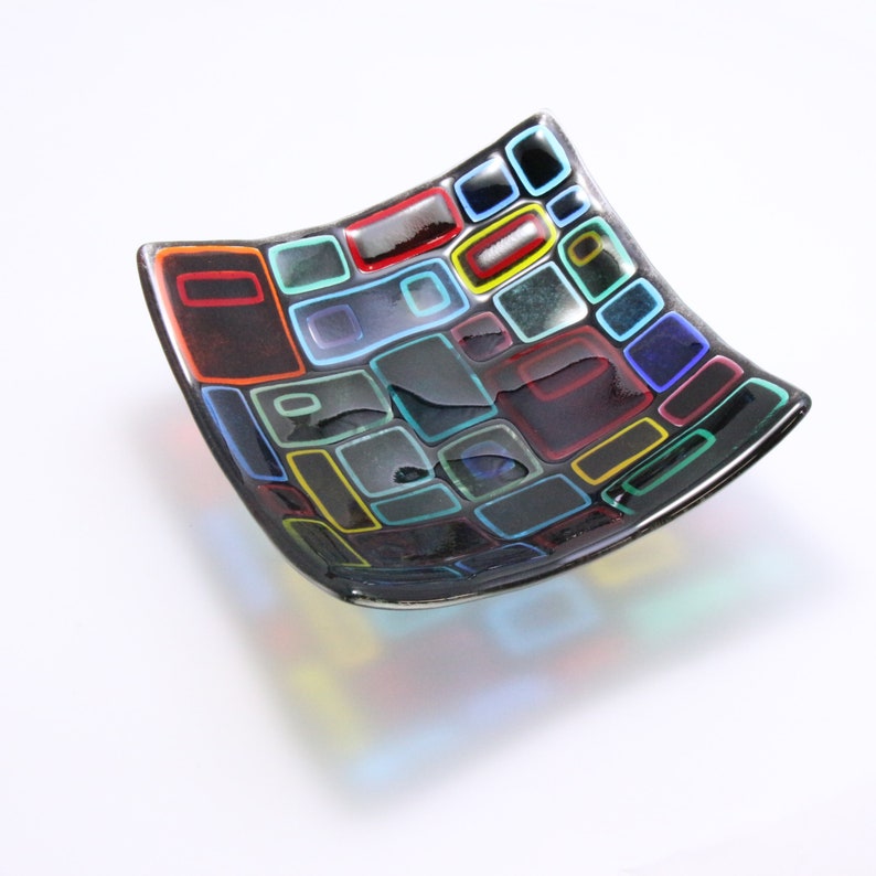 Puede incluir: Un plato cuadrado de vidrio con fondo negro y dise&ntilde;os geom&eacute;tricos coloridos. El plato est&aacute; hecho de vidrio y tiene un acabado brillante.