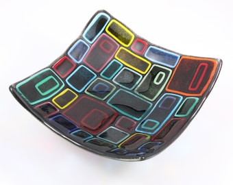 May include: A square glass dish with a black background and a colorful geometric pattern of squares and rectangles. The squares and rectangles are outlined in black and filled with various colors, including red, yellow, blue, green, and orange.