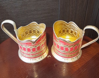 May include: Two gold-colored metal shot glasses with red and gold decorative patterns. The glasses have a scalloped rim and a handle.