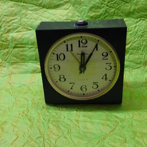Könnte beinhalten: Ein schwarzer Wecker mit weißem Zifferblatt und schwarzen Zeigern. Die Uhr hat einen goldenen Rand und die Zahlen 1 bis 12 sind in Schwarz gedruckt. Die Uhr ist auf 11:55 eingestellt.