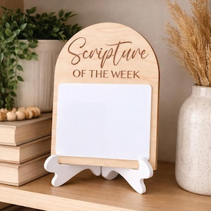 May include: Wooden sign with the words "Scripture of the Week" engraved at the top. The sign has a white rectangular area for writing and is supported by a white stand. The sign is made of light-colored wood.