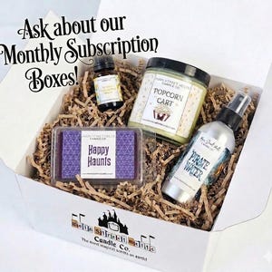 May include: A white gift box filled with brown paper crinkle filler containing a small amber glass bottle, a jar candle labelled Popcorn Cart, a rectangular wax melt labelled Happy Haunts, and a spray bottle labelled Pirate Water. Text reads Ask about our Monthly Subscription Boxes!