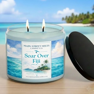 May include: A light blue two-wick candle in a glass jar with a black lid. The label features a tropical ocean scene with palm trees and the text: Main Street Melts Candle Co. Soar Over Fiji Prepare For Takeoff www.MainStreetMeltsCandleCo.com.