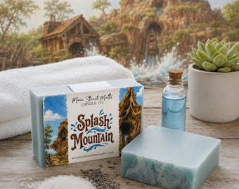 May include: A rectangular bar of light blue soap with a label reading Main Street Melts Candle Co. Splash Mountain, alongside a second plain light blue soap bar, a small glass bottle with a cork stopper, white bath salts, and dried lavender.