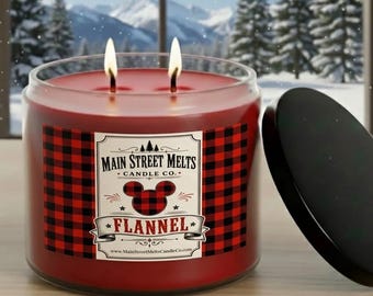 May include: A red two-wick candle in a glass jar with a black lid. The label features a red and black buffalo plaid pattern, a mouse ear silhouette, and the text Main Street Melts Candle Co. Flannel with a website address at the bottom.