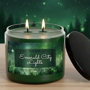 May include: A dark green two-wick candle in a glass jar with a black lid. The label features a forest and starry sky graphic with the text Main Street Melts Candle Co, Emerald City Lights, and www.MainStreetMeltsCandleCo.com in white script.