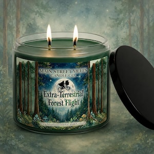 May include: A dark green two-wick candle in a clear glass jar with a black lid. The label features a forest scene with a full moon, a silhouette of two figures on a bicycle, and the text Main Street Melts Candle Co. Extra-Terrestrial Forest Flight.