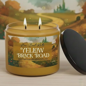 May include: A yellow candle in a glass jar with two lit wicks and a black lid. The label features a landscape illustration and the text Main Street Melts YELLOW BRICK ROAD www.MainStreetMeltsCandleCo.com. The wax is a golden yellow colour.
