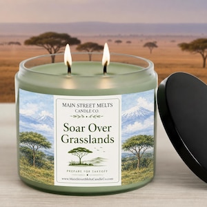 May include: A two-wick candle in a clear glass jar with a sage green wax fill. The label features a watercolour landscape of mountains and acacia trees with the text Main Street Melts Candle Co. Soar Over Grasslands Prepare For Takeoff.