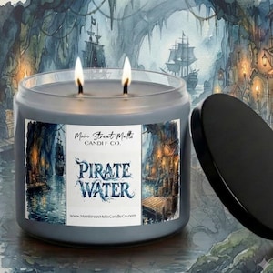 Peut inclure: Une bougie grise à deux mèches dans un pot en verre avec un couvercle noir. L'étiquette présente une illustration à l'aquarelle d'un navire pirate dans une grotte et le texte Main Street Melts CANDLE CO. PIRATE WATER et www.MainStreetMeltsCandleCo.com.