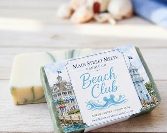 Könnte beinhalten: Ein rechteckiges Stück Seife, umwickelt mit einem Papieretikett, das eine Aquarellillustration eines Küstengebäudes und eines Leuchtturms zeigt. Der Text lautet Main Street Melts Candle Co. Beach Club, Green Clover, Crisp Aloe und die Webadresse.