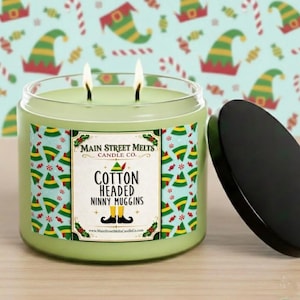 May include: A light green two-wick candle in a glass jar with a black lid. The label features a repeating pattern of elf hats and the text Main Street Melts Candle Co. Cotton Headed Ninny Muggins with an illustration of elf shoes and holly.