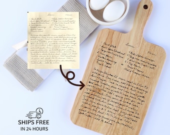 May include: A wooden cutting board with a handle, engraved with a handwritten recipe. The board is light brown and features a list of ingredients and instructions. Two white eggs sit in a small white bowl. A beige towel is also in the image.