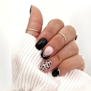 Può includere: Una mano con unghie curate con vari design. Le unghie sono dipinte di nero, rosa e con stampa leopardata. Alcune unghie hanno dettagli argentati. La mano indossa anelli d'argento e poggia su un tessuto bianco a coste.