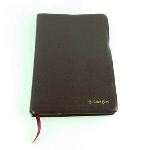 May include: A dark brown leather-bound journal with a gold cross and the name "Karen Clay" embossed on the front cover. A burgundy ribbon bookmark is attached to the spine.
