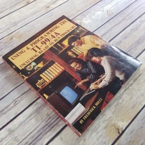 May include: A red and white book titled "Using & Programming the TI-99/4A Including Ready-to-Run Programs" by Frederick Holtz. The cover features a photo of three people using a computer.