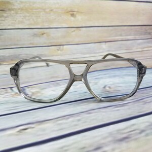 May include: Clear aviator-style eyeglasses with a gray frame. The glasses have a double bridge and rectangular temples. The lenses are clear, and the frame is a translucent gray color. The glasses are set against a light wood background.
