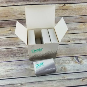 May include: A box of Deter Deodorant Soap. The box is open and shows three bars of soap inside. A single bar of soap is sitting on a wooden surface. The soap is gray with green lettering that reads "Deter Deodorant Soap" and "Net Wt 3.25 oz (92 g)".