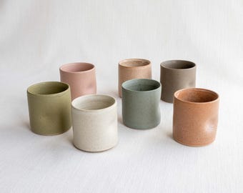 May include: A collection of seven ceramic cups in various muted earth tones including sage green, dusty pink, beige, light gray, dark taupe, and terracotta. Each cylindrical cup features a smooth matte finish and a subtle indentation on the side.